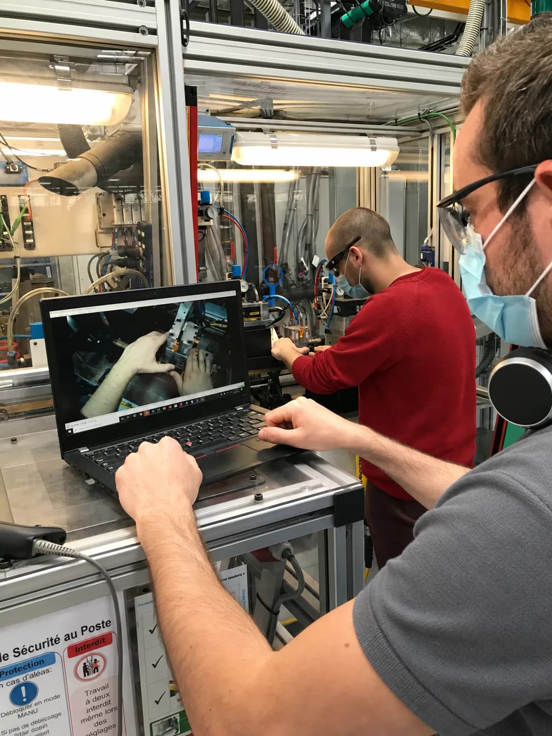 An operator works on a machine while a colleague reviews a video on a laptop